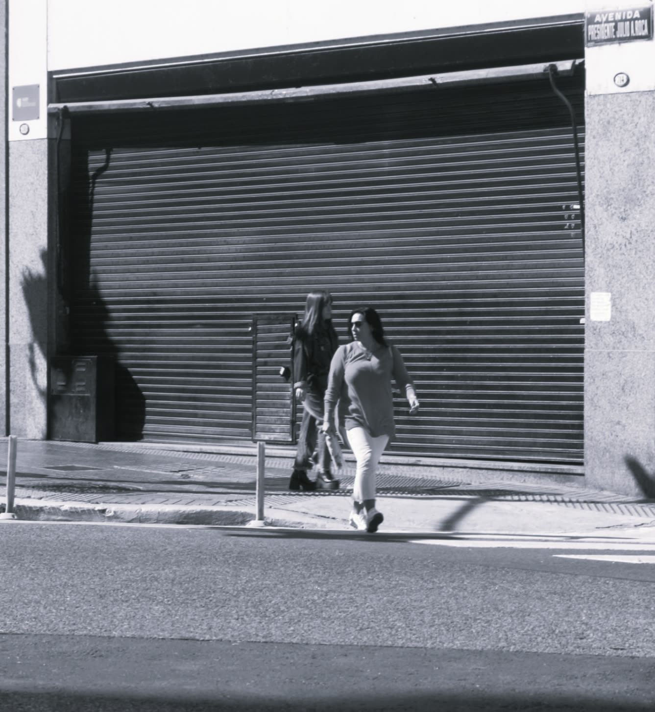Dos personas caminando junto a una persiana metálica, blanco y negro